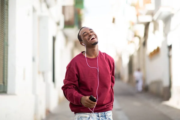 街の外を歩く携帯電話やイヤホンを持つ笑顔の若い黒人男性の肖像画 — ストック写真
