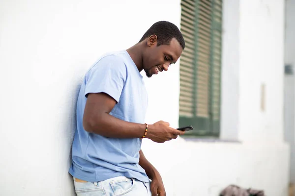 携帯電話を見ながら外の白い壁に傾いているハンサムな若いアフリカ人男性のサイドポートレート — ストック写真