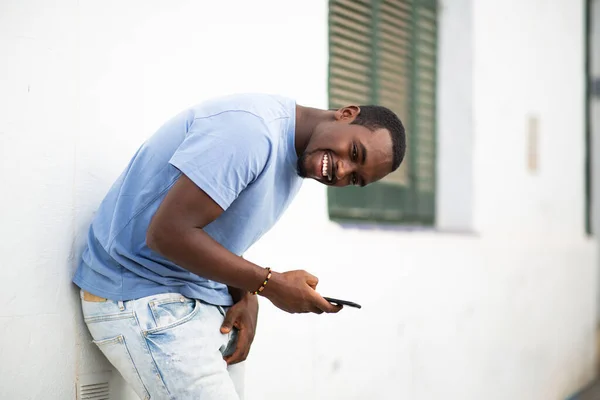 携帯電話が外の壁に傾いている陽気な若いアフリカの男の肖像画 — ストック写真