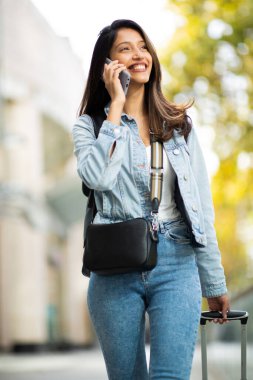 Şehirde cep telefonu ve çantasıyla yürüyen gülümseyen seyahat eden kadının portresi.