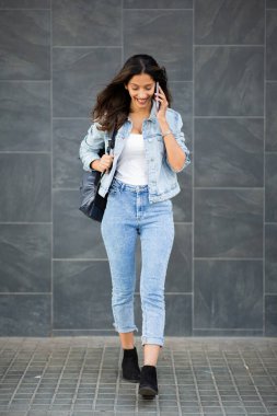 Duvarda cep telefonuyla yürüyen gülümseyen genç bir kadının tam boy portresi