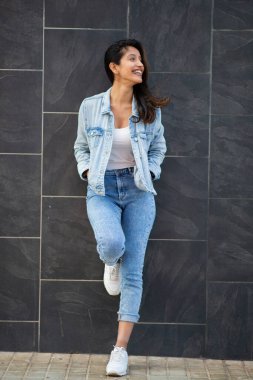 Duvara yaslanmış gülümseyen genç bir kadının tam boy portresi.