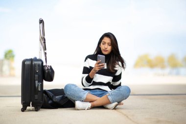 Tüm vücut portresi. Genç seyahat eden kadın. Cep telefonu ve bavuluyla yerde oturuyor.