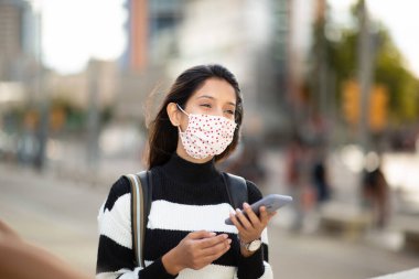 Dışarıda cep telefonuyla duran yüz maskeli genç bir kadının portresi. 