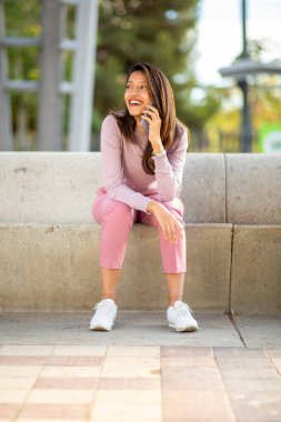 Dışarıda oturmuş cep telefonuyla konuşan gülümseyen bir kadının tam boy portresi. 