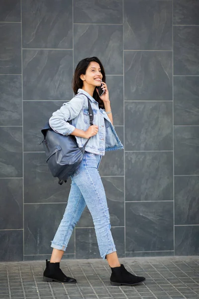 携帯電話と話を壁で歩く幸せな女性の完全な長辺の肖像画 — ストック写真