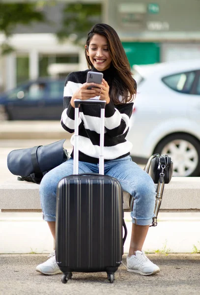 全身人像笑容满面的旅行女孩坐在城市里 手里拿着手机和手提箱 — 图库照片