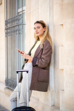 Dış bina duvarına seyahat çantasıyla yaslanırken cep telefonu kullanan ve başka yere bakan bir kadının portresi