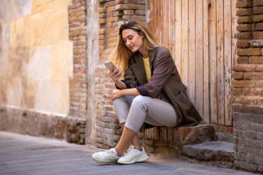 Güzel beyaz kadın dışarıda eski ahşap kapıya karşı otururken cep telefonuyla mesajlaşıyor.