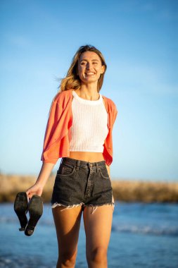 Sahilde yürüyen şortlu mutlu genç bir kadının portresi.