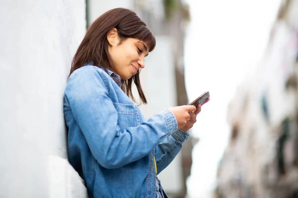 年轻漂亮的拉丁女人靠在墙上看手机的侧像 — 图库照片