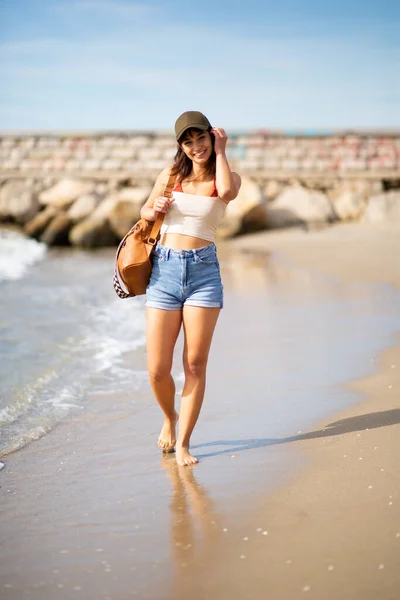 头戴帽子 提着袋子在海滩上行走的年轻貌美女子的全身像 — 图库照片