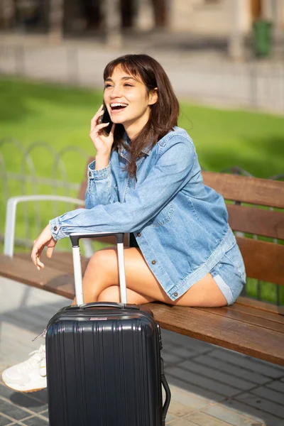 一个微笑的年轻女性旅行者的画像 她坐在外面的长椅上 提着手提箱 用手机交谈 — 图库照片