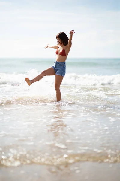 ビーチで水を蹴る興奮した若い女性の完全な長さの肖像画 — ストック写真