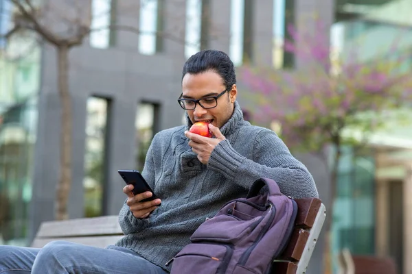 市内のベンチの外に座っている間 若いアフリカ人男性がリンゴを食べて携帯電話を使っている姿 — ストック写真