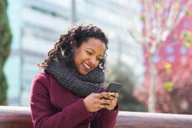 Dışarıda oturan ve cep telefonu kullanan kışlık montlu ve eşarplı güzel siyah kadın portresi.