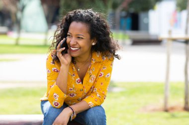 Dışarıda oturan ve cep telefonuyla konuşan güzel siyahi bir kadının portresini kapat.