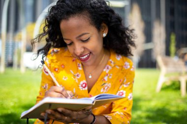 Parkta oturmuş kitap yazan mutlu Afrikalı genç bir kadının portresini yakından çek.