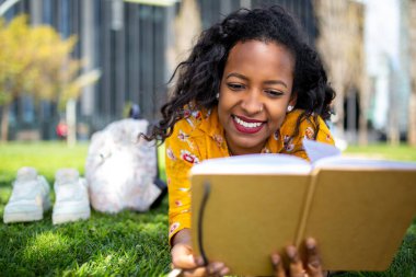 Parkta çimlere uzanmış kitap okuyan mutlu Afrikalı genç bir kadının portresini yakından çek.