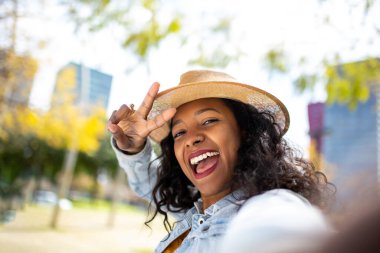 Neşeli, Afro-Amerikalı, şapkalı, selfie çeken ve dışarıda barış işareti gösteren genç bir kadının portresini yakından çek.