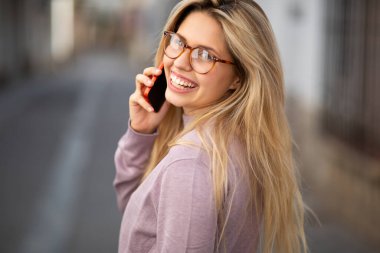 Gözlüklü, cep telefonuyla konuşan ve dışarıda yürürken arkasına bakan neşeli genç bir kadının arka plan portresi.