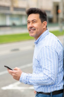 Şehirde cep telefonuyla dolaşan olgun bir adamın yan portresi.