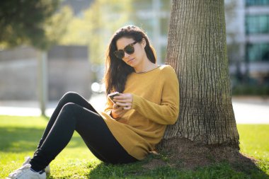 Güneş gözlüğü takan güzel Asyalı kadının portresi. Parktaki ağacın yanında otururken akıllı telefondan mesaj atıyor.