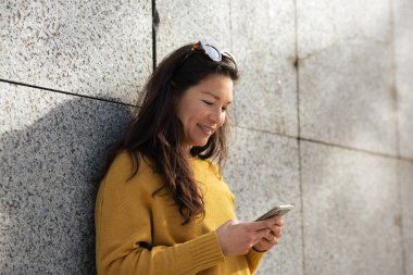 Duvara yaslanmış ve dışarıda cep telefonu kullanan mutlu Asyalı kadın portresi.