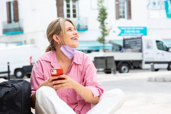 面带微笑的年轻女子坐在外面 带着面具和手机 望着远方的画像 — 图库照片