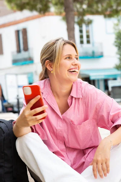 一个快乐的年轻女子坐在外面 拿着手机 望着别处 笑着的画像 — 图库照片