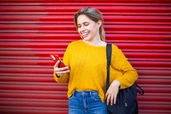 赤いシャッターの前に立っている間に彼女の携帯電話と笑顔を見て幸せな白人女性の肖像画 — ストック写真