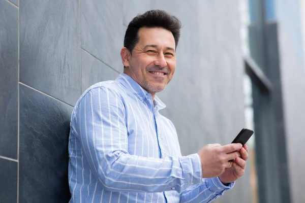Portret Uśmiechniętego Dojrzałego Latynoskiego Mężczyzny Opierającego Się Ścianę Telefonem Komórkowym — Zdjęcie stockowe