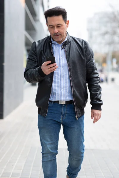 Portrait of stylish older latin man in black leather jacket walking outside looking at cellphone