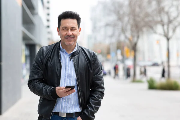 Portrait of handsome mature latin man wearing leather jacket with smartphone standing outside in the city