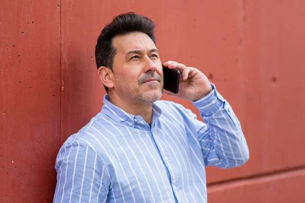 赤い壁に寄りかかって携帯電話で話している思慮深い成熟した男のクローズアップ肖像画 — ストック写真