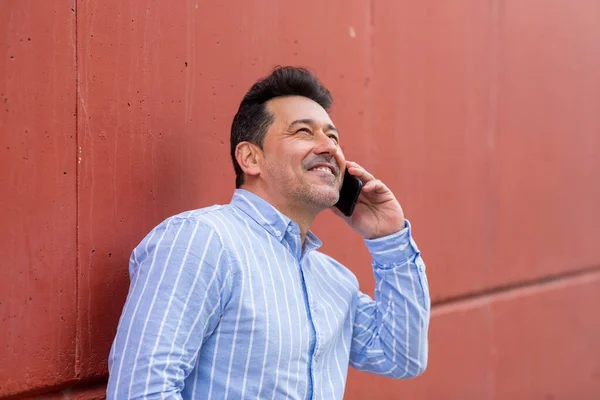 幸せな成熟した男の肖像画は赤い壁に傾いて 携帯電話で話しています — ストック写真