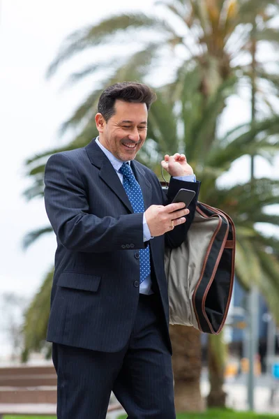 スーツの旅行袋で屋外に歩いて彼の携帯電話を使用して成熟したビジネスマンの笑顔の肖像画 — ストック写真