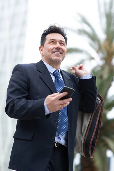 スーツバッグと外を歩く携帯電話を持つハンサムな成熟したビジネスマンの肖像画 — ストック写真