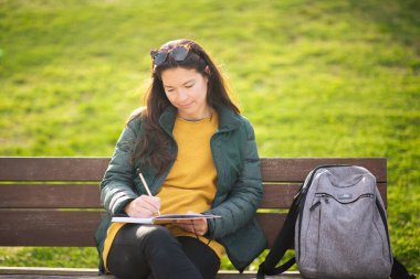 Bankta oturan genç bir kadının portresi. Dışarıda bir parkta deftere notlar yazıyor.