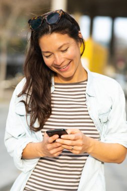 Şık Asyalı kadın portresi. Akıllı telefonuyla mesaj atıyor. Şehrin dışında duruyor.