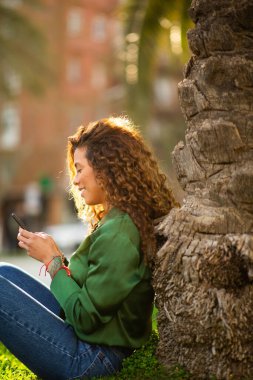 Şehir parkındaki bir ağaca yaslanmış akıllı telefon kullanan İspanyol kadının yan portresi.
