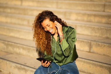 Şehirde merdivenlerde oturmuş kulaklık ve cep telefonuyla müzik dinleyen gülümseyen Latin kadının portresi.
