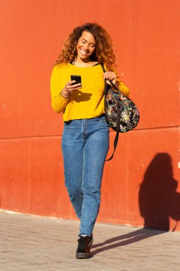 Dışarıda çantalı ve cep telefonlu gülümseyen genç Latin kadının vücut portresi.