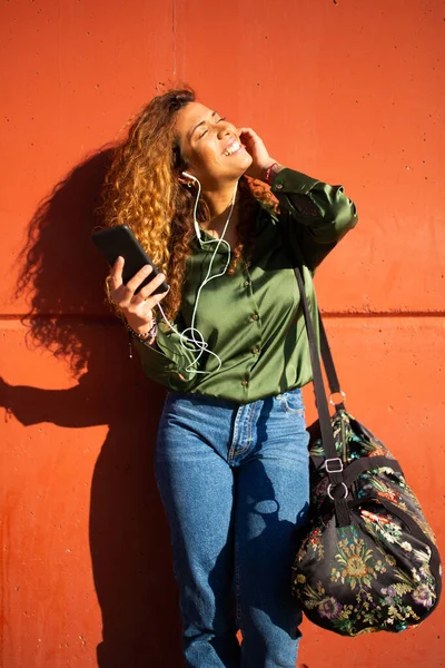 明るい若いラテン語の女性の肖像 スマートフォンで音楽を聴いてオレンジの壁に傾いて — ストック写真