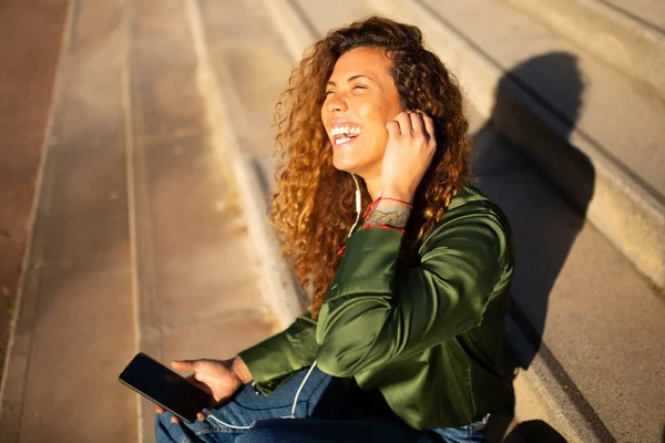 市内の階段の外に座っている間 イヤホンや携帯電話で音楽を聞いて楽しむ陽気な女性の肖像画 — ストック写真