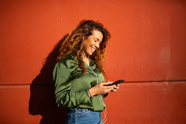 美しい若いラテン語の女性の肖像イヤフォンや携帯電話で音楽を聞いてオレンジの壁にもたれて — ストック写真