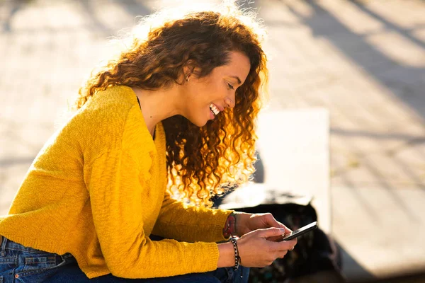 陽気な若い女性の携帯電話でテキストメッセージのサイドポートレート市内の外に座って — ストック写真