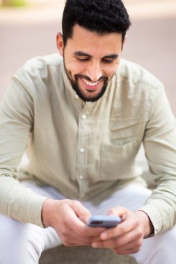 Dışarıda oturmuş cep telefonundan mesaj okuyan gülümseyen genç adamın portresi.