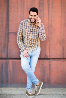 Kahverengi duvarın önünde cep telefonuyla konuşan gülümseyen genç Arap 'ın tam boy portresi