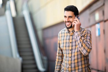 Gülümseyen genç Arap adamın portresi. Dışarıda yürüyor ve cep telefonuyla konuşuyor.